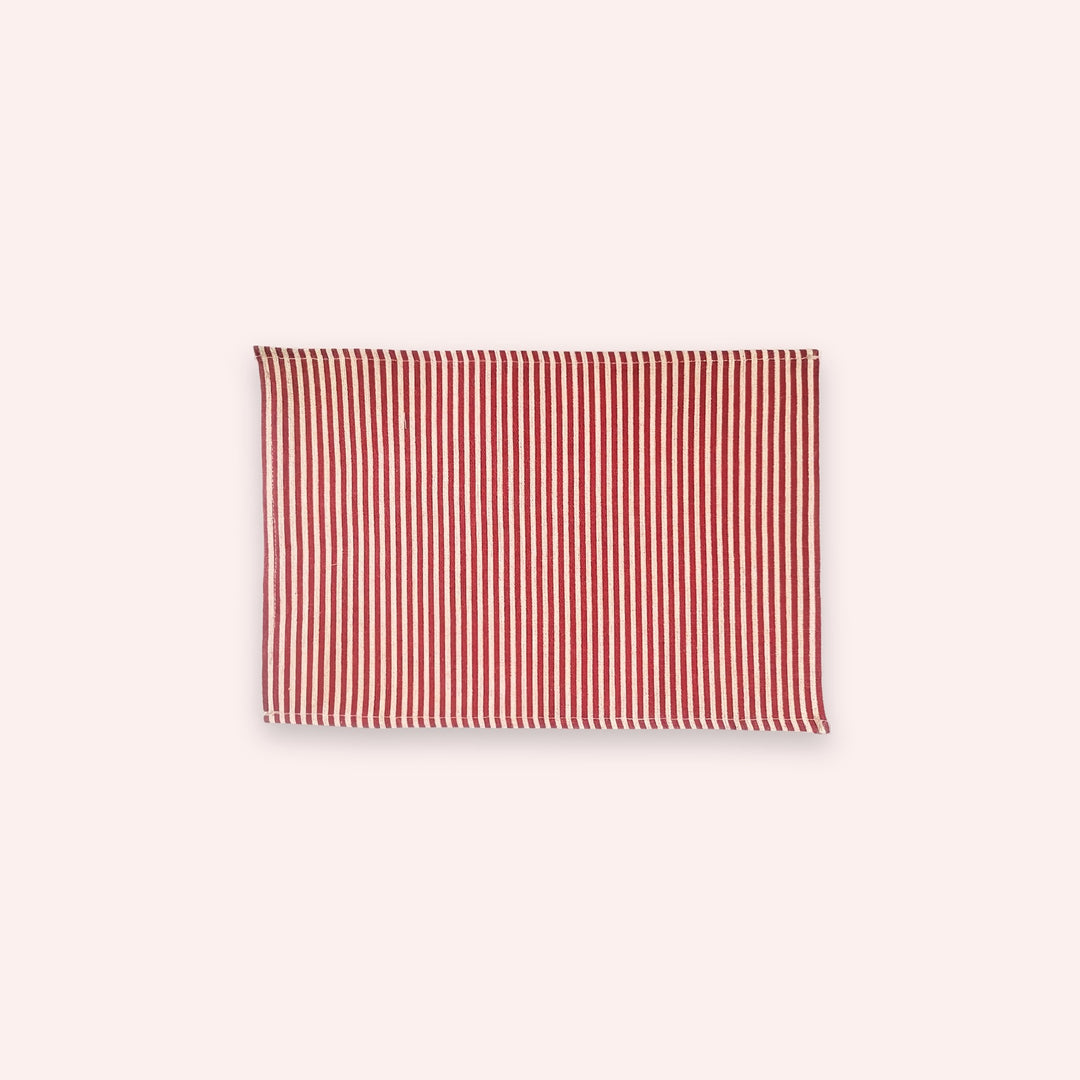A rectangular fabric item, likely a placemat or small towel, featuring a vertical stripe pattern in red and off-white, centered on a solid pale pink background.