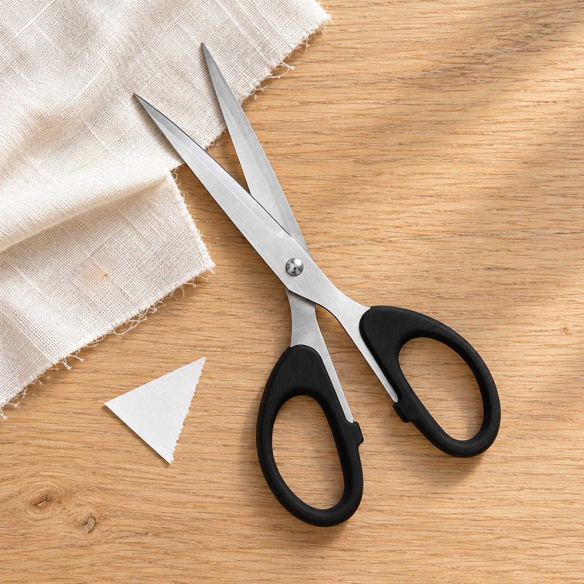 Black-handled stainless steel scissors open beside a partly cut cotton swatch and a paper offcut on a pale oak craft table
