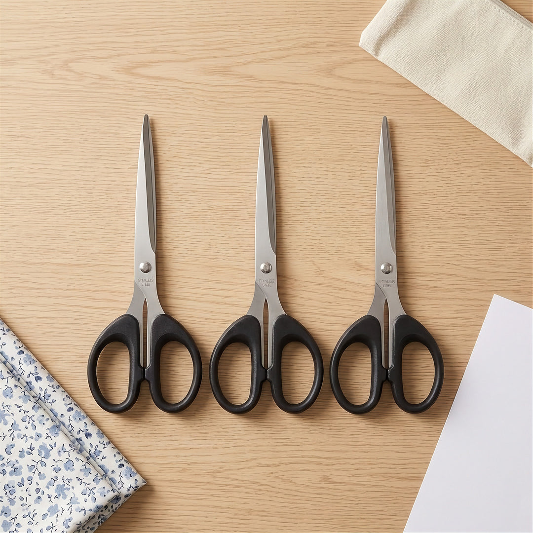 Three pairs of scissors with black handles and silver blades are arranged side by side on a light wood tabletop, with fabric and paper props partially visible around them.
