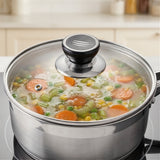 A stainless steel pot on a stovetop with a clear glass lid and black knob, showing cooked vegetables like carrots, peas, and celery in broth.