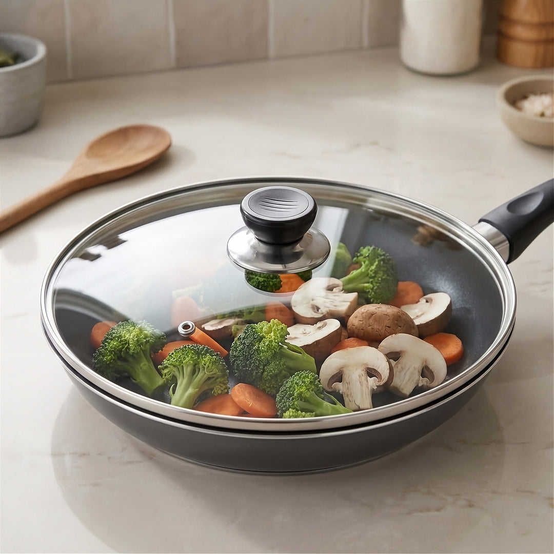 A dark gray non-stick frying pan with a clear glass lid and black handle, shown on a kitchen countertop with broccoli, carrots, mushrooms, and a wooden spoon nearby.