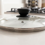 A close-up of a glass cooking lid with a black knob and stainless steel rim, resting on a light countertop with a blurred kitchen background.