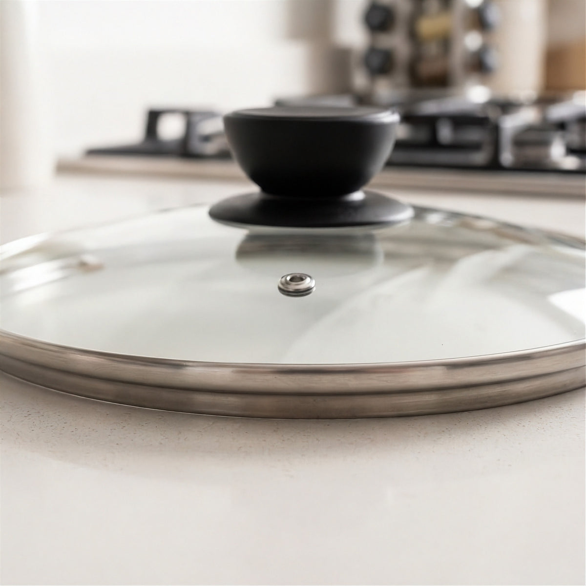 A close-up of a glass cooking lid with a black knob and stainless steel rim, resting on a light countertop with a blurred kitchen background.