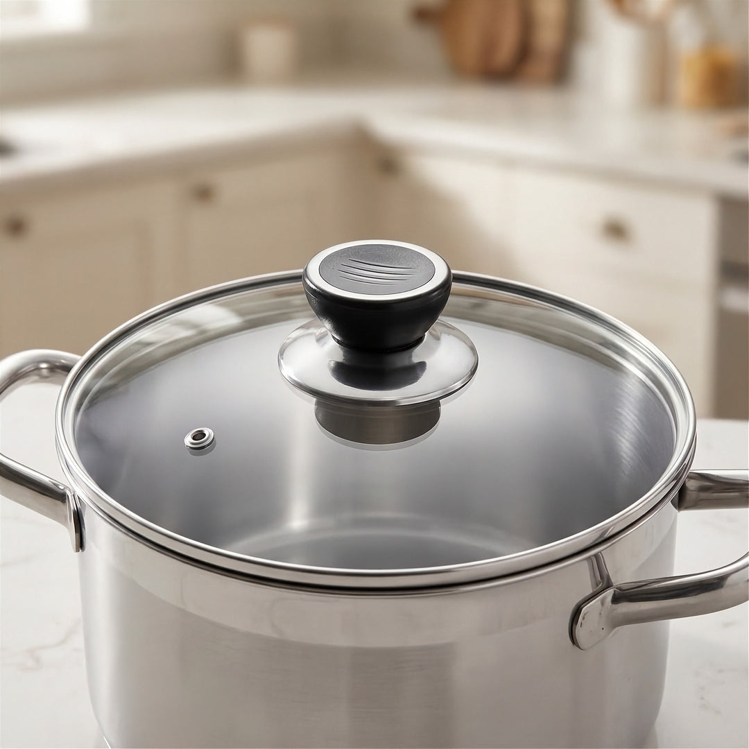 A stainless steel pot with a clear glass lid and a black knob, shown on a kitchen countertop with a blurred kitchen background.
