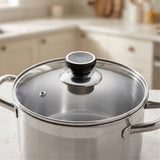 A stainless steel pot with a clear glass lid and a black knob, shown on a kitchen countertop with a blurred kitchen background.