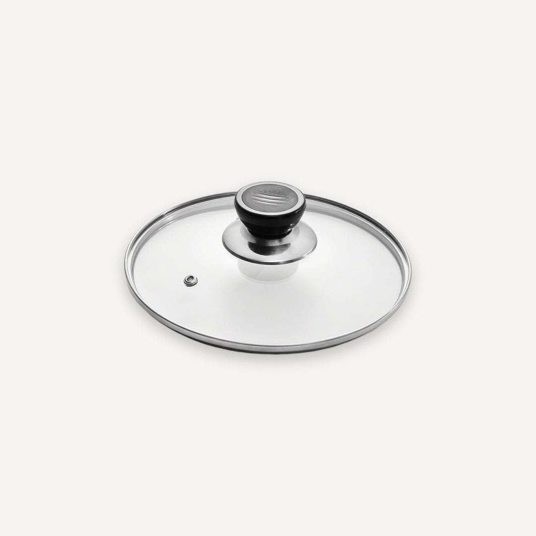 A clear glass cookware lid with a brushed silver metal rim and a black-and-silver knob, shown on a clean off-white background.