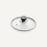 A clear glass cookware lid with a brushed silver metal rim and a black-and-silver knob, shown on a clean off-white background.