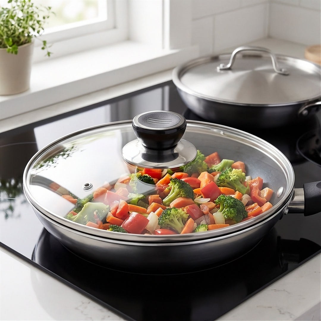 A black non-stick frying pan with a clear glass lid sits on a black induction cooktop, cooking a colorful mix of broccoli, carrots, and red peppers. A matching black pan with a stainless steel lid is visible in the background on a white tiled kitchen counter.