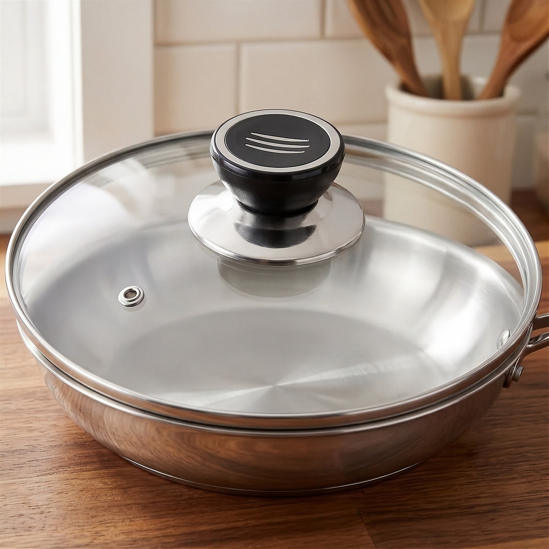 A stainless steel pan with a clear glass lid and a black knob, shown on a wooden countertop in a kitchen setting.