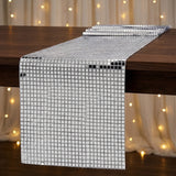 A long rectangular table runner made of small reflective silver mirror tiles, draped over a dark wooden table with warm string lights blurred in the background.