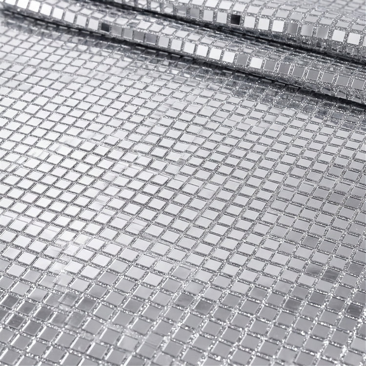 A close-up of a silver mirrored mesh fabric with small square reflective tiles, shown folded to reveal its texture and reflective properties.