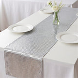 A silver table runner with a small square mosaic pattern laid down the center of a white tablecloth, styled with white plates and a small vase of white flowers.