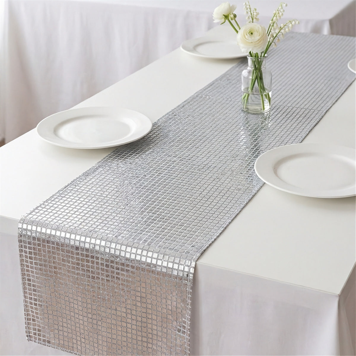 A silver table runner with a small square mosaic pattern laid down the center of a white tablecloth, styled with white plates and a small vase of white flowers.