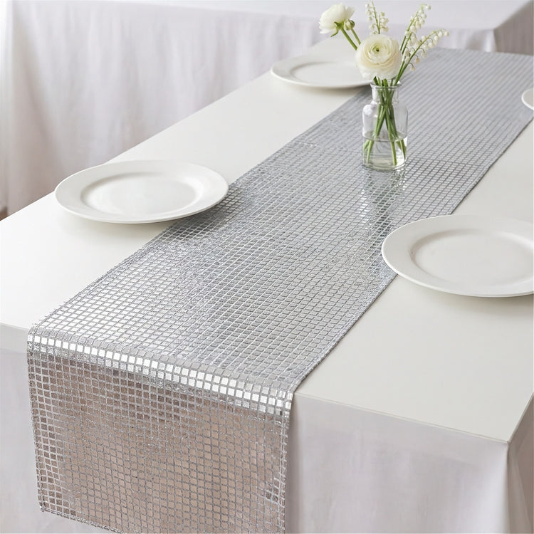 A silver table runner with a small square mosaic pattern laid down the center of a white tablecloth, styled with white plates and a small vase of white flowers.