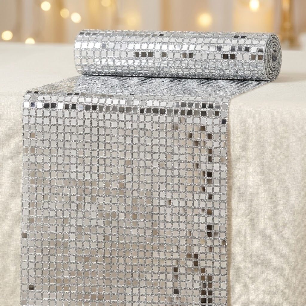 A roll of silver mirrored table runners with a grid of small square reflective tiles, shown partially unrolled on a light beige tablecloth with warm blurred lights in the background.