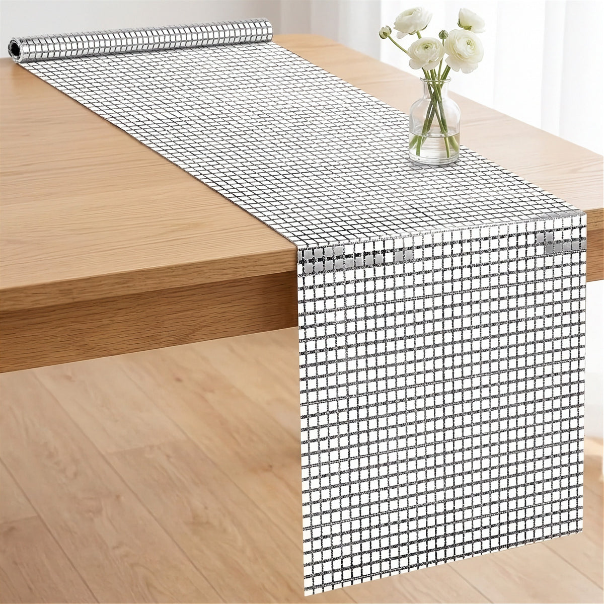 A silver and white mosaic table runner with a reflective rhinestone-like grid pattern, shown draped over a light wood table with a small vase of white flowers on top.