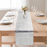 A rectangular table runner with a shimmering silver mosaic tile pattern, styled on a light wood dining table with white plates, wine glasses, and a small vase of flowers.