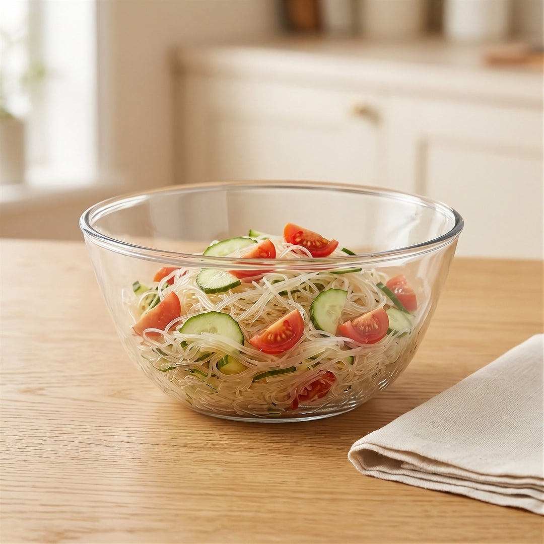 A clear glass salad bowl filled with noodles, cucumber slices, and tomato wedges, set on a light wood table with a folded cloth napkin and a softly blurred kitchen background.
