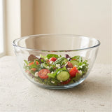 A clear glass salad bowl filled with mixed greens, tomatoes, cucumbers, and feta cheese, set on a light stone surface near a window.
