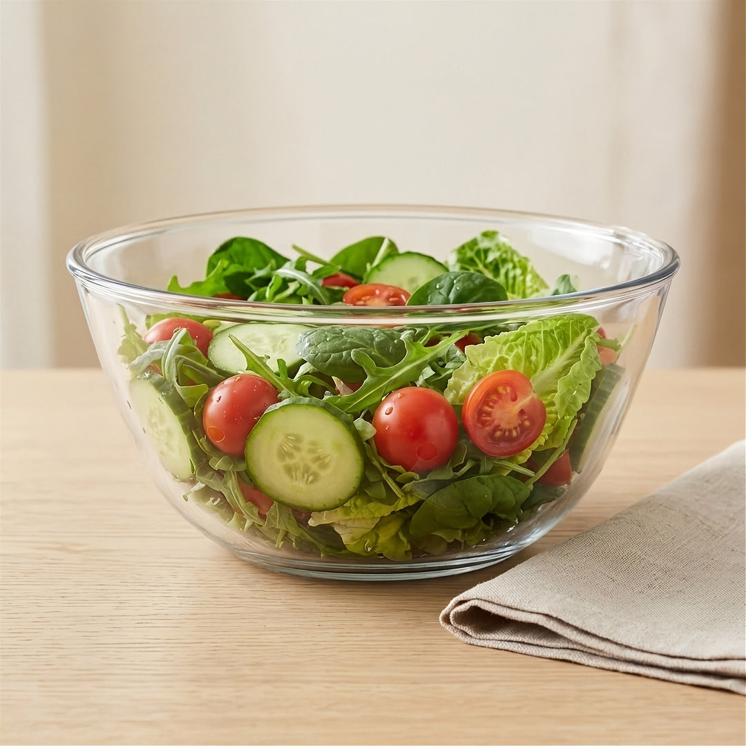 A clear glass salad bowl filled with mixed greens, cucumber slices, and cherry tomatoes on a light wood table, with a folded beige napkin beside it.