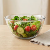 A clear glass salad bowl filled with mixed greens, cucumber slices, and cherry tomatoes on a light wood table, with a folded beige napkin beside it.