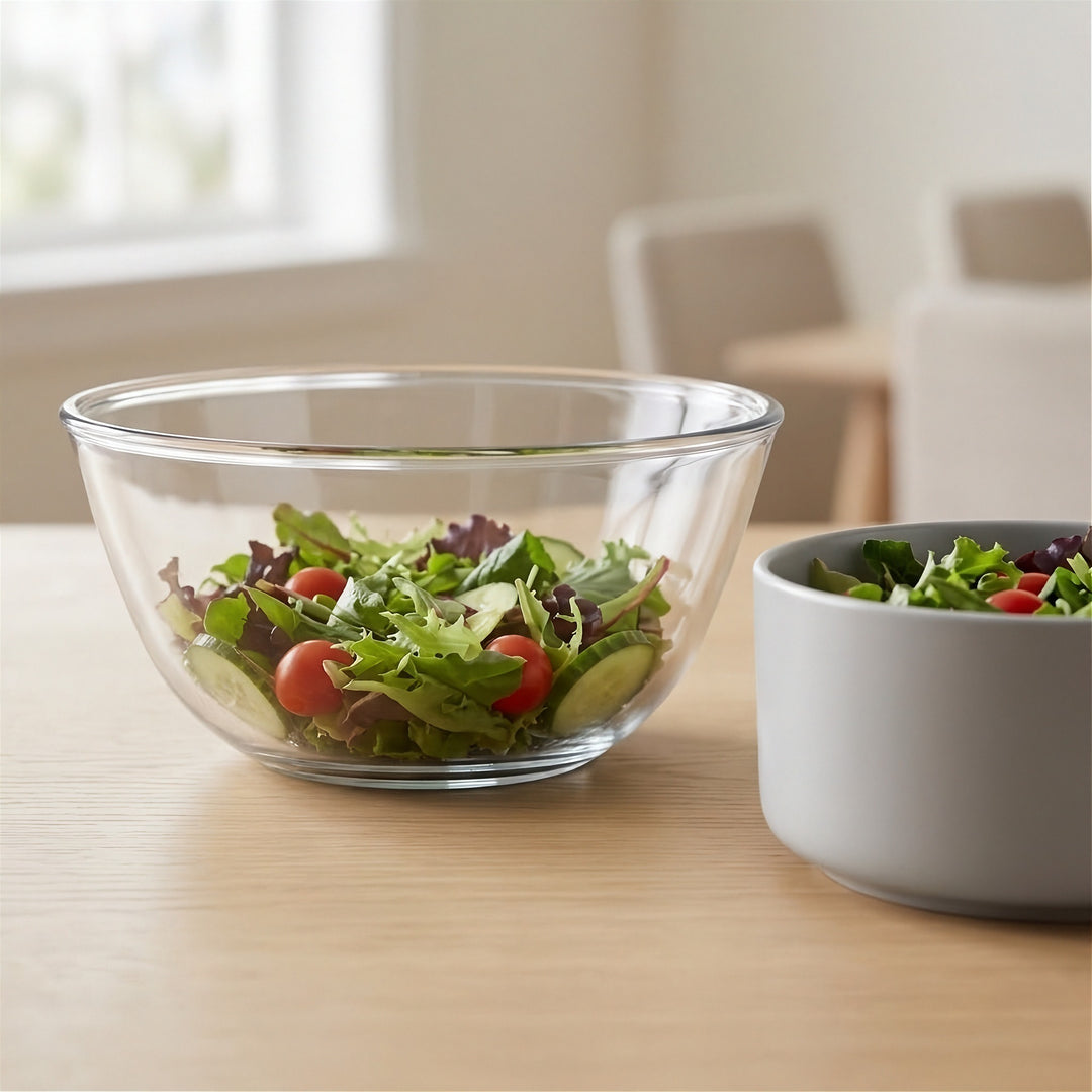 A clear glass salad bowl filled with mixed greens and cherry tomatoes sits on a light wood table, with a gray ceramic bowl partially visible at right in a softly blurred indoor setting.