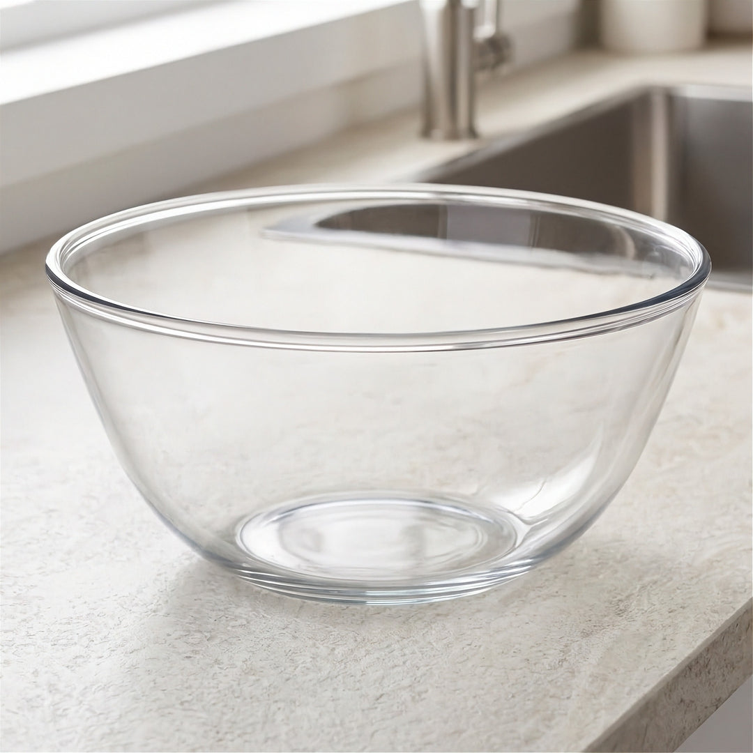 A clear glass mixing bowl with a wide rim, placed on a light beige kitchen countertop near a sink.