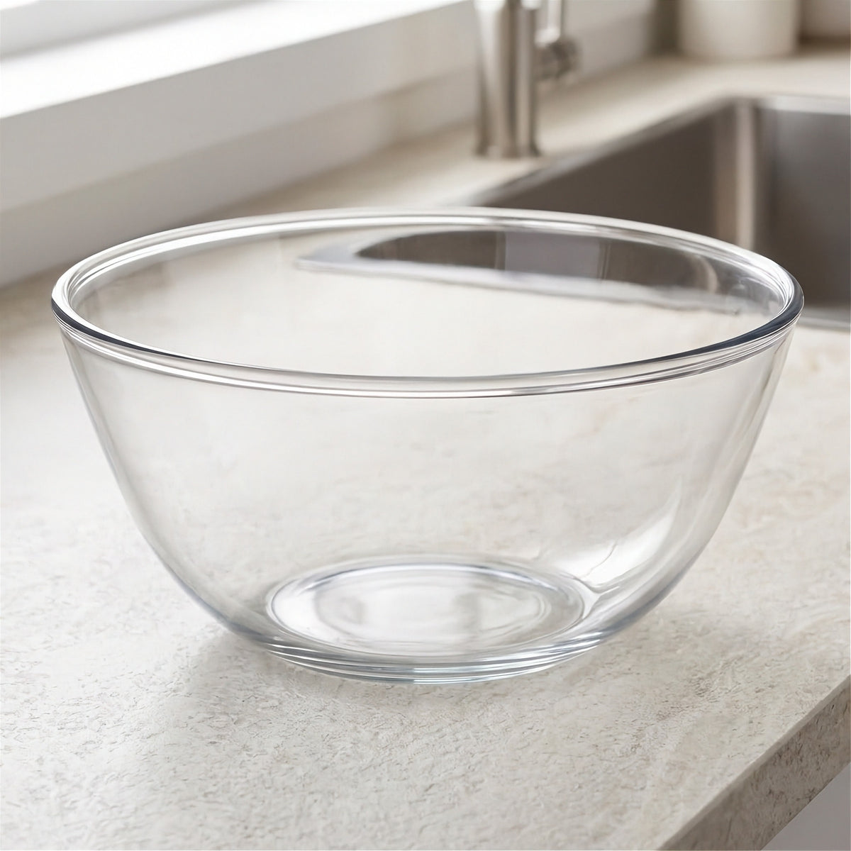 A clear glass mixing bowl with a wide rim, placed on a light beige kitchen countertop near a sink.