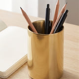 A brushed gold metal pencil holder cup on a light wood desk, holding black pens and wooden pencils next to an open notebook.