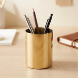 A shiny gold cylindrical desk organizer cup filled with pens and a pencil, placed on a light wood desk with a blurred notebook in the background.