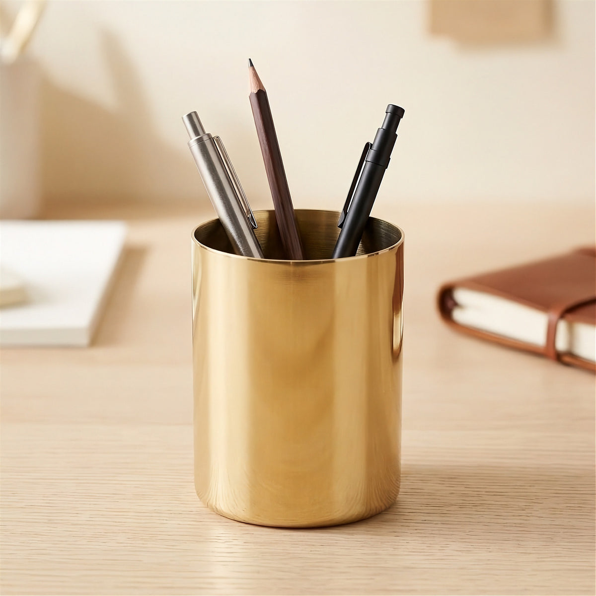 A shiny gold cylindrical desk organizer cup filled with pens and a pencil, placed on a light wood desk with a blurred notebook in the background.