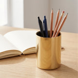 A shiny gold metal pencil cup filled with pens and pencils, sitting on a light wood desk next to an open notebook.