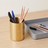 A brushed gold metal pencil and pen holder cup sitting on a light wood desk next to a gray tray.