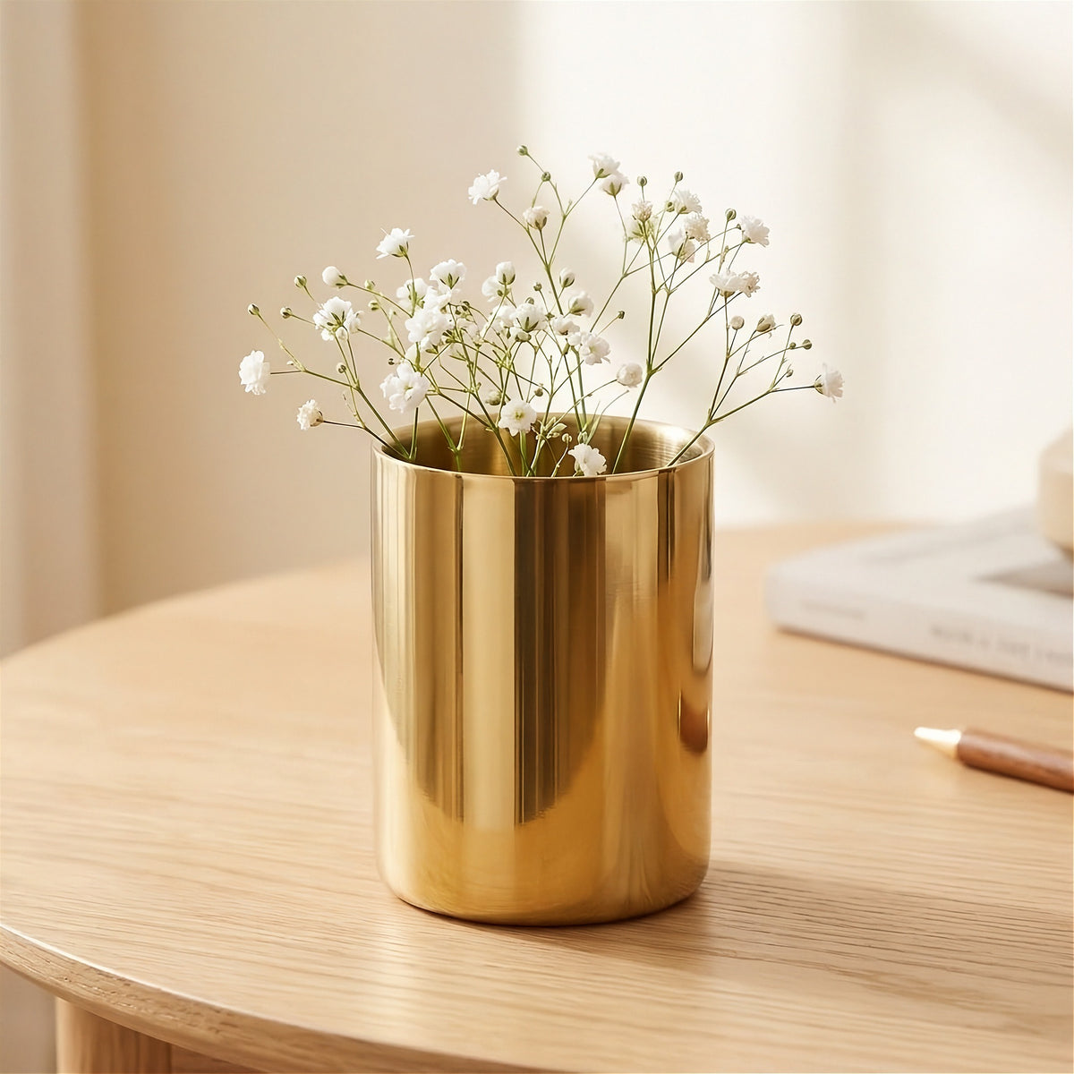 A shiny gold cylindrical vase filled with small white baby's breath flowers, placed on a light wood table in a softly lit indoor setting.