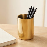 A shiny gold cylindrical pen holder on a light wood desk, holding four black pens, with a neutral beige background and a white notebook corner visible.
