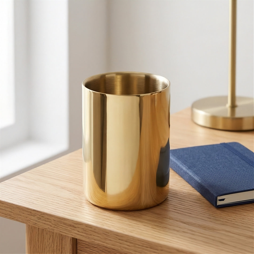 A shiny gold cylindrical cup or tumbler sits on a light wood table next to a blue notebook, with a softly blurred lamp and window in the background.