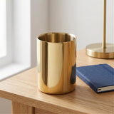 A shiny gold cylindrical cup or tumbler sits on a light wood table next to a blue notebook, with a softly blurred lamp and window in the background.