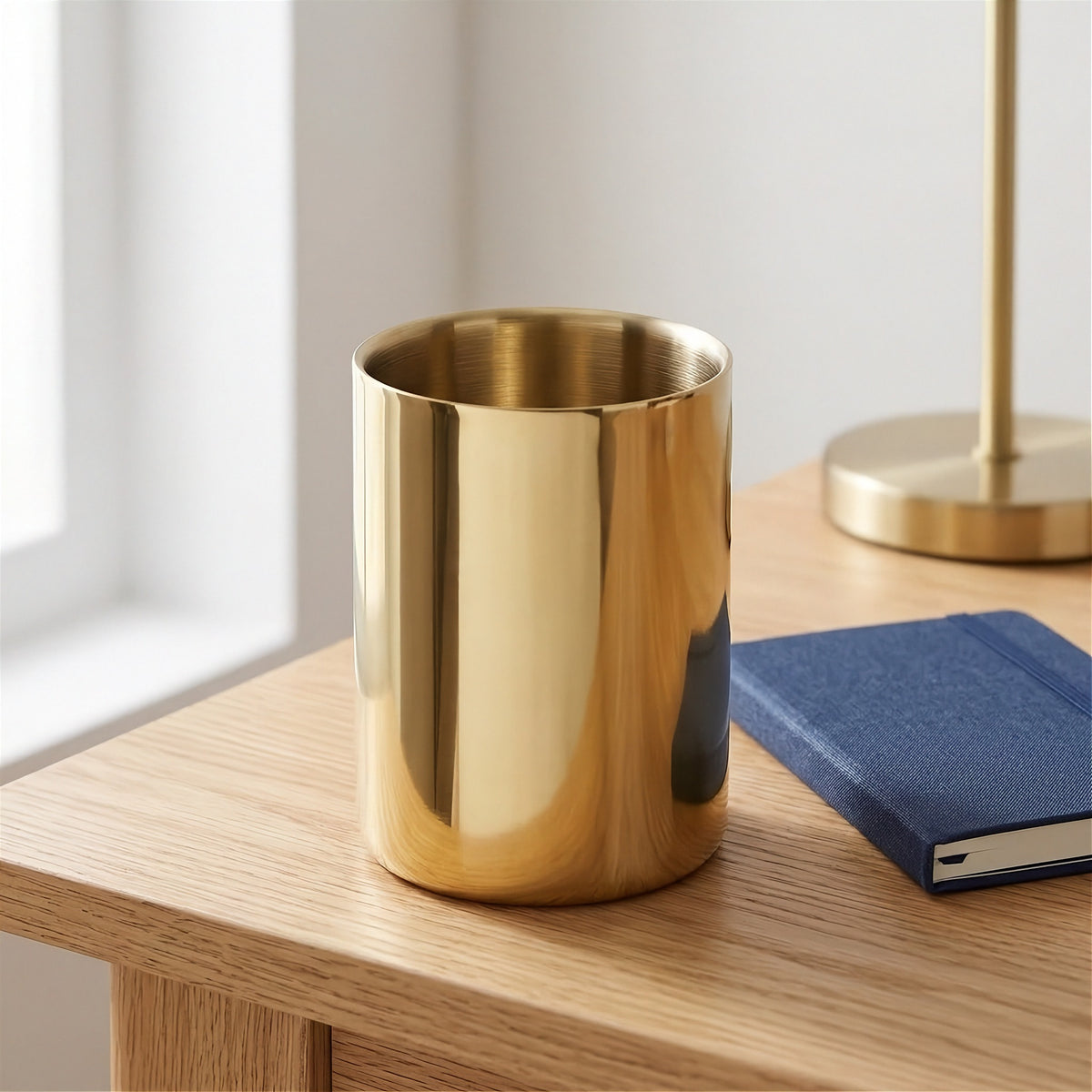A shiny gold cylindrical cup or tumbler sits on a light wood table next to a blue notebook, with a softly blurred lamp and window in the background.