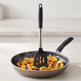 A silicone spatula with a black handle and a stainless steel shaft is resting inside a frying pan filled with roasted potatoes and carrots on a light countertop.