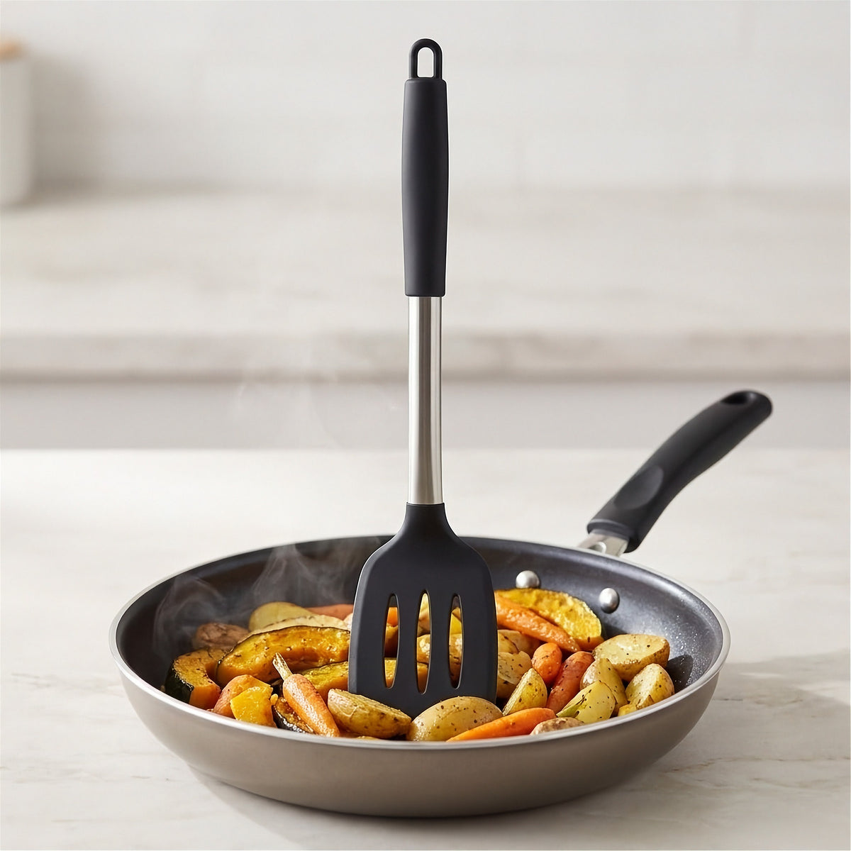 A silicone spatula with a black handle and a stainless steel shaft is resting inside a frying pan filled with roasted potatoes and carrots on a light countertop.