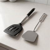 Two kitchen utensils on a light countertop: a black silicone spatula with a silver handle and a stainless steel scraper with a loop handle.