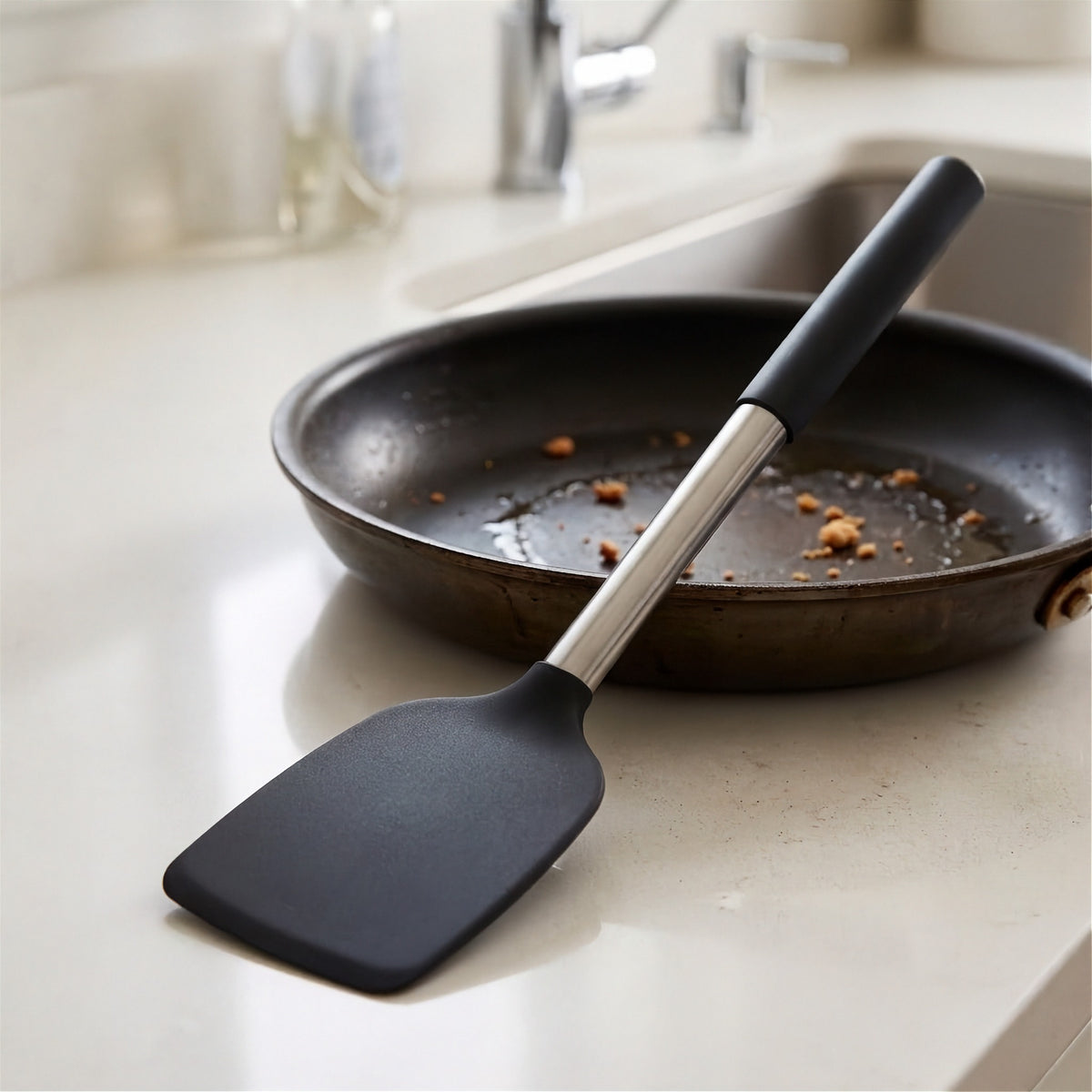 A black silicone spatula with a silver metal handle rests on a light beige kitchen countertop next to a dark non-stick frying pan.
