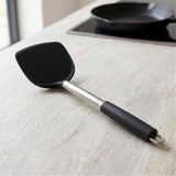 A black silicone spatula with a stainless steel shaft and black handle grip, resting on a light beige stone countertop with a stovetop pan blurred in the background.