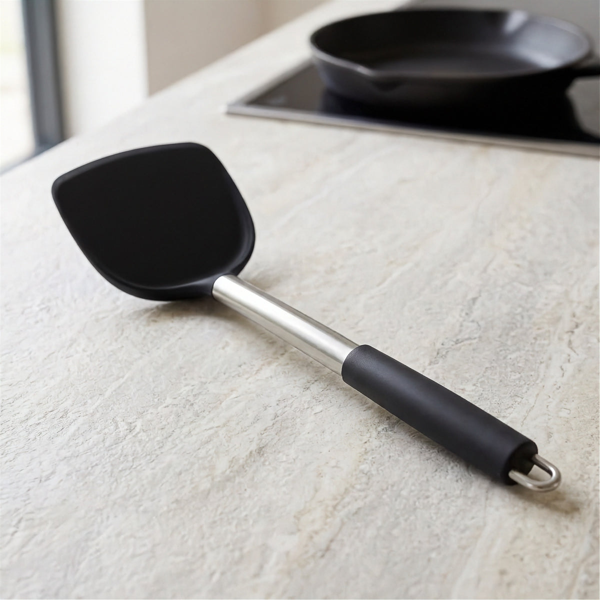 A black silicone spatula with a stainless steel shaft and black handle grip, resting on a light beige stone countertop with a stovetop pan blurred in the background.