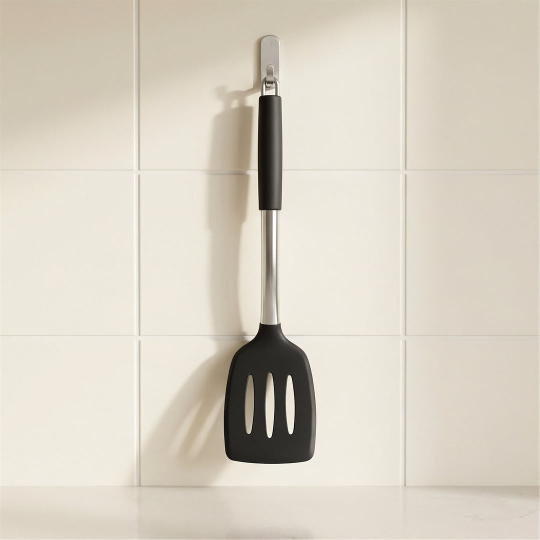 A black silicone spatula with a stainless steel handle and silver wall hook, hanging on a white tiled wall.