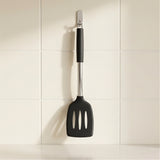 A black silicone spatula with a stainless steel handle and silver wall hook, hanging on a white tiled wall.