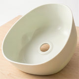 A modern, asymmetrical ceramic sink basin in a light cream color, shown from an elevated angle on a light wood surface.