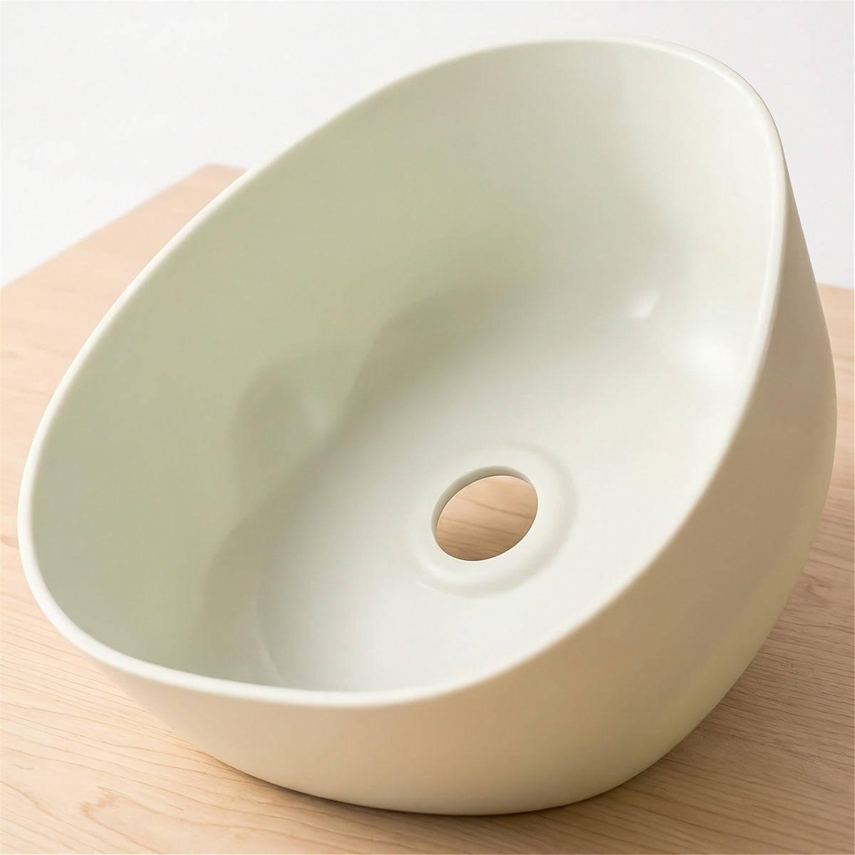 A modern, asymmetrical ceramic sink basin in a light cream color, shown from an elevated angle on a light wood surface.