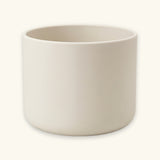 A minimalist, matte ceramic cylindrical planter in a warm off-white or cream color, shown against a matching light cream background.