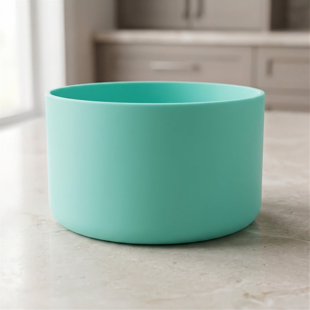 A matte mint green cylindrical bowl or planter sitting on a light beige marble countertop with a blurred kitchen background.